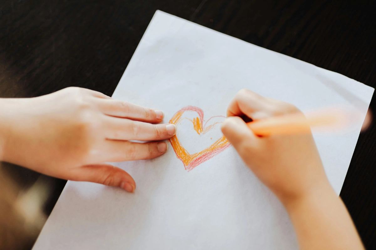 Close up of a child’s hands drawing a red and orange colored heart on white paper