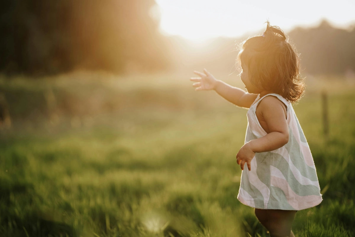 A young girl standing in a field in a green and pink dress with her arm outstretched as the sun sets in the background