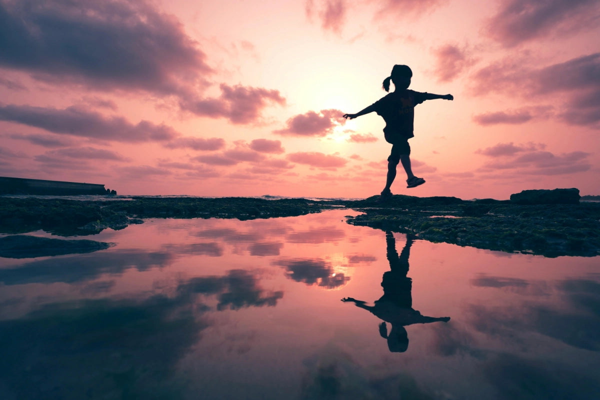 A silhouette of a child skipping along rocks around a lake with water reflecting the pink sunset